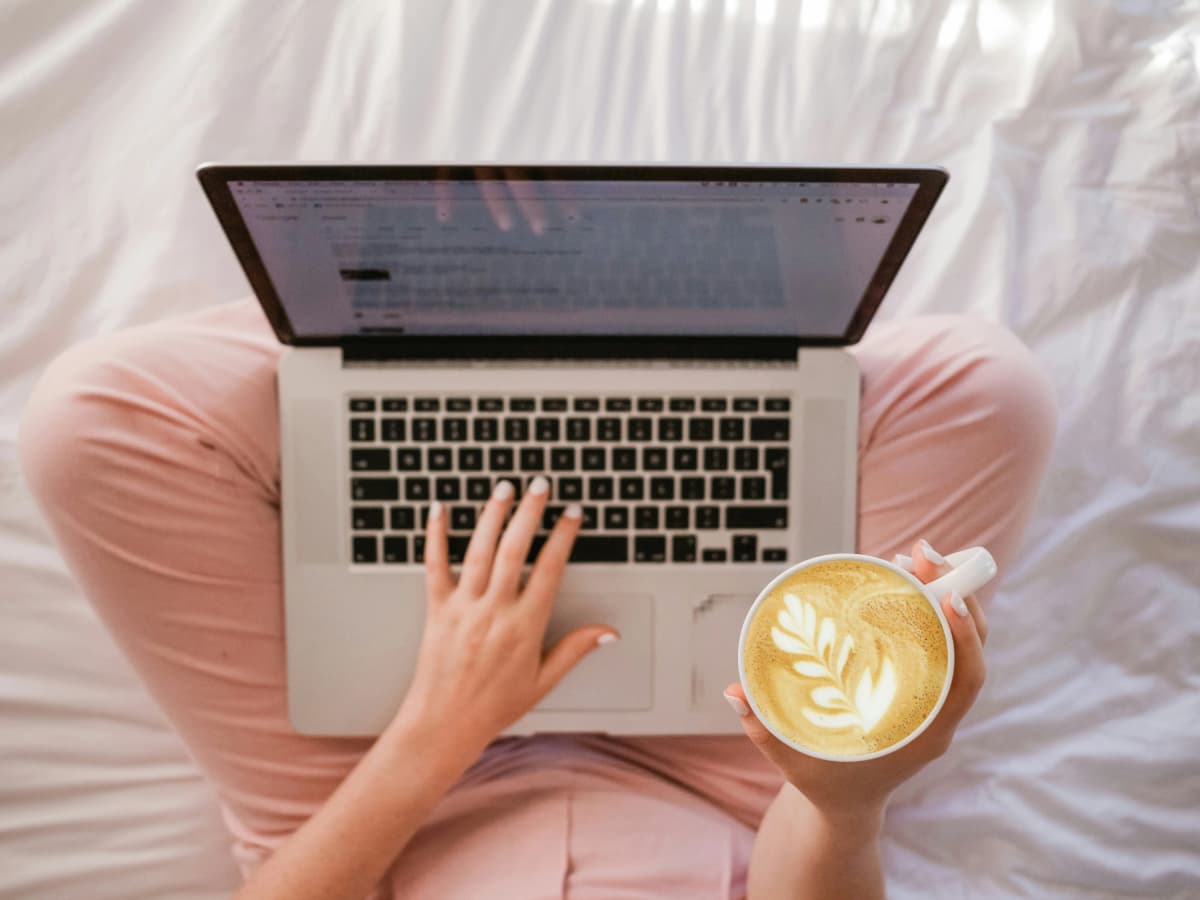 A girl sitting with her laptop, wearing pink trousers and holding a coffee on her right hand.